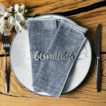 Gümüş Bismillah yazılı metal peçete süsü, mavi keten peçeteli tabakta, çatal ve bıçakla