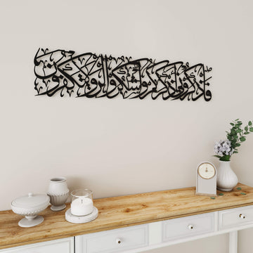Bakara 152. Ayet Şükür ve Zikir uzun hat metal tablo, siyah renk, açık duvarda konsol ve vazo yanında orta mesafeden görünüm