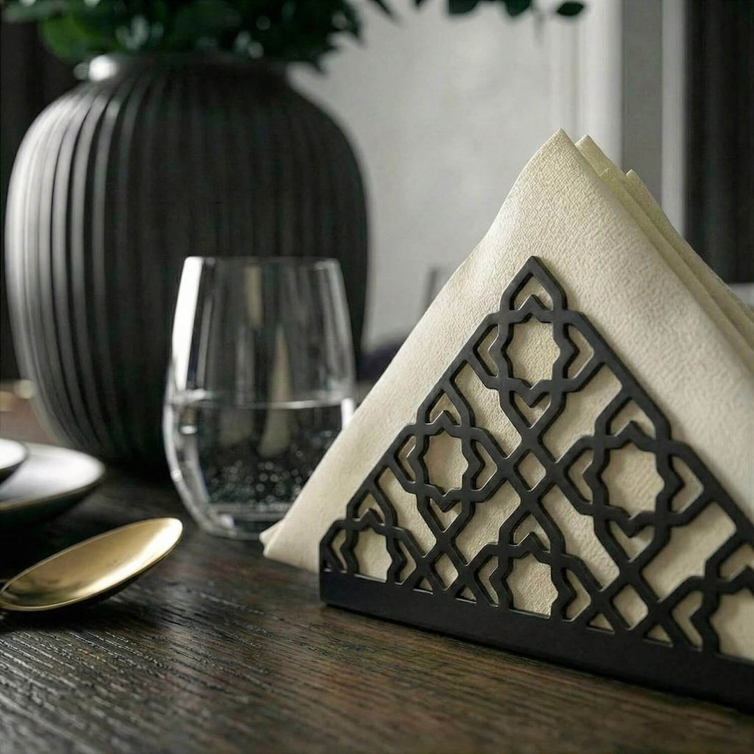 Geometrik desenli siyah metal peçetelik, koyu ahşap yemek masasında bardak yanında yakın kadraj
