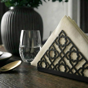 Geometrik desenli siyah metal peçetelik, koyu ahşap yemek masasında bardak yanında yakın kadraj