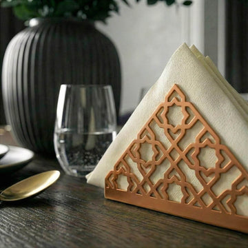 Geometrik desenli bakır metal peçetelik, koyu ahşap yemek masasında bardak yanında yakın kadraj