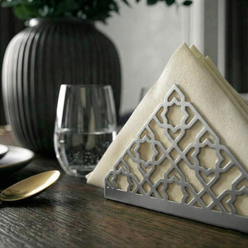 Geometrik desenli gümüş metal peçetelik, koyu ahşap yemek masasında bardak yanında yakın kadraj