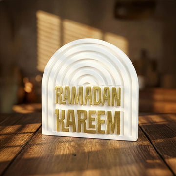 Altın yaldızlı Ramazan Kerim yazılı kemerli beyaz taş dekor, ahşap masada ışık gölge altında ön görünüm