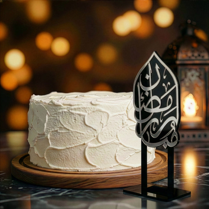 beyaz kremayla kapli yuvarlak pastanin yaninda siyah ayakli gumus renkli arapca ramazan kerim yazili akrilik kek topper arka planda sicak tonlu isiklar ve yanan dekoratif fener ile estetik ramazan sunumu islamicwallarttr