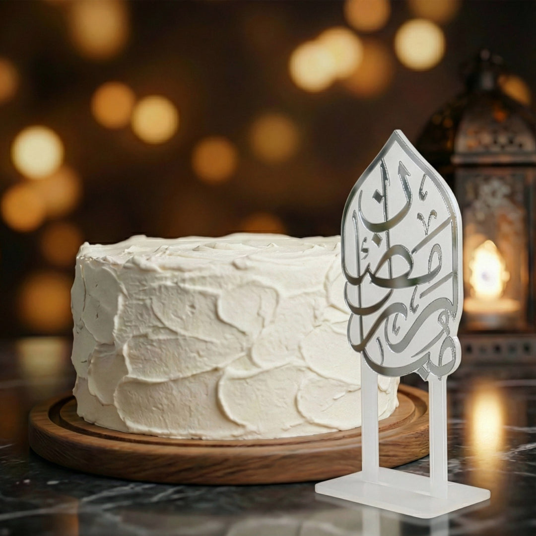 beyaz kremayla kapli yuvarlak pastanin yaninda ayakli gumus renkli arapca ramazan kerim yazili akrilik kek topper arka planda sicak tonlu isiklar ve yanan dekoratif fener ile estetik ramazan sunumu islamicwallarttr