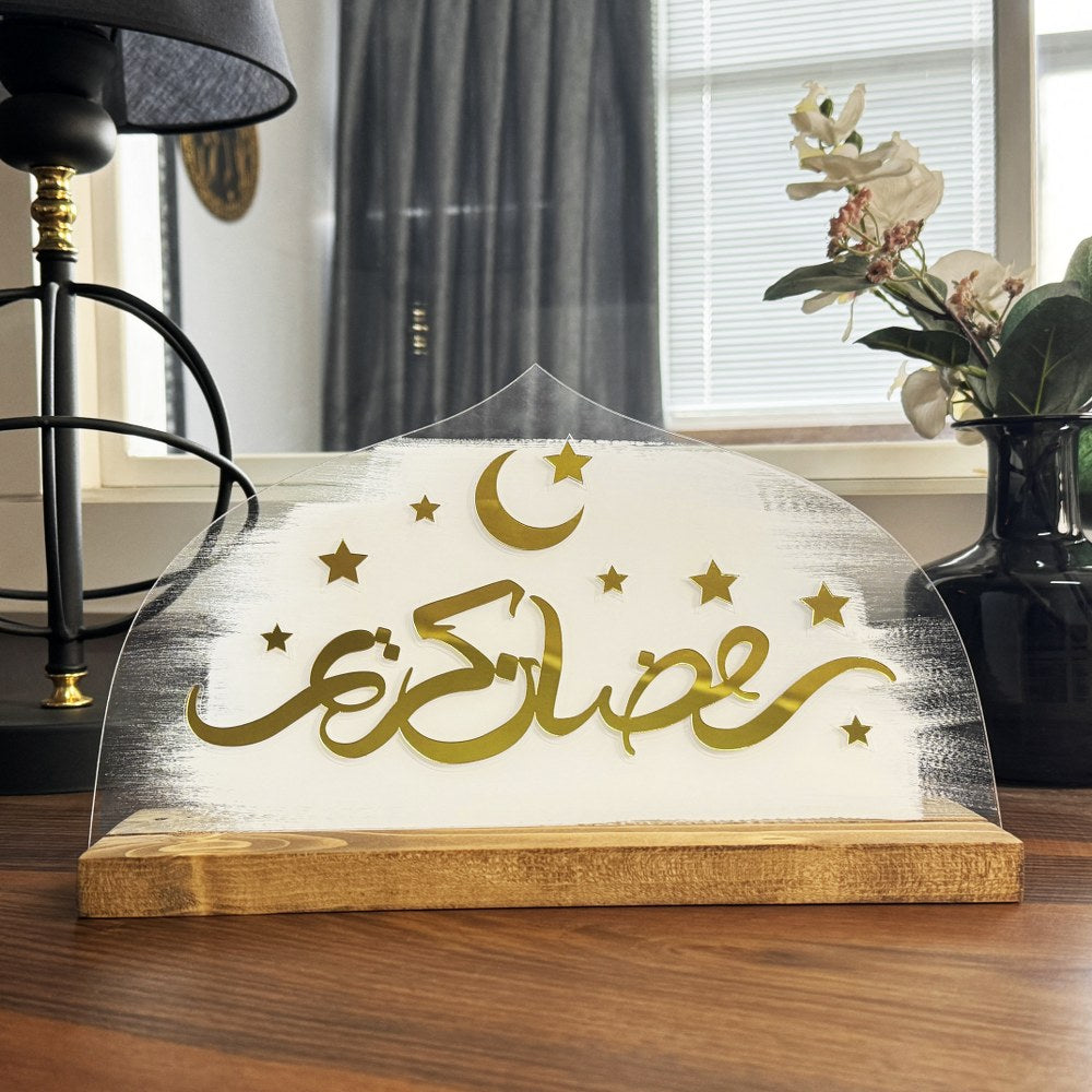 Beyaz fırça darbeli pleksi üzerinde altın Ramazan Kerim yazısı, ahşap stand ve pencere önünde yakın görünüm
